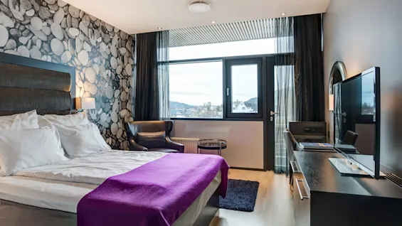 Et moderne hotellrom har en seng med et lilla teppe, en steinmønstret vegg, en lenestol, en TV og vinduer med fjellutsikt.