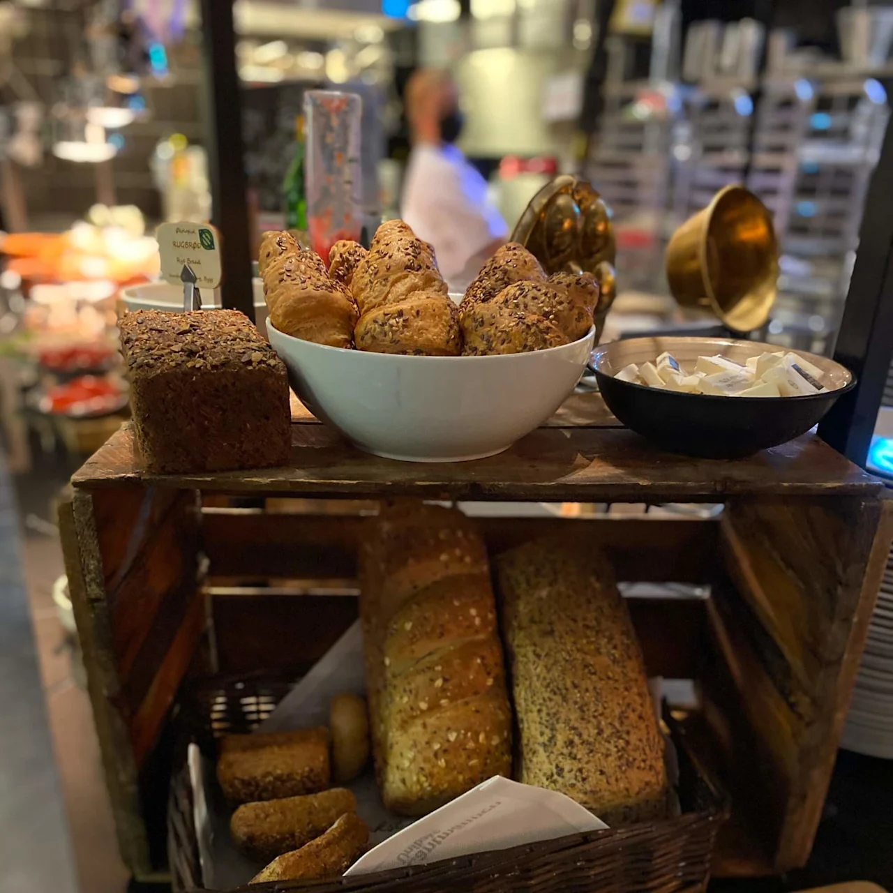 A wooden shelf displays various breads, including seeded rolls and loaves, alongside butter packets. A busy kitchen environment is visible in the background, with trays and a chef.