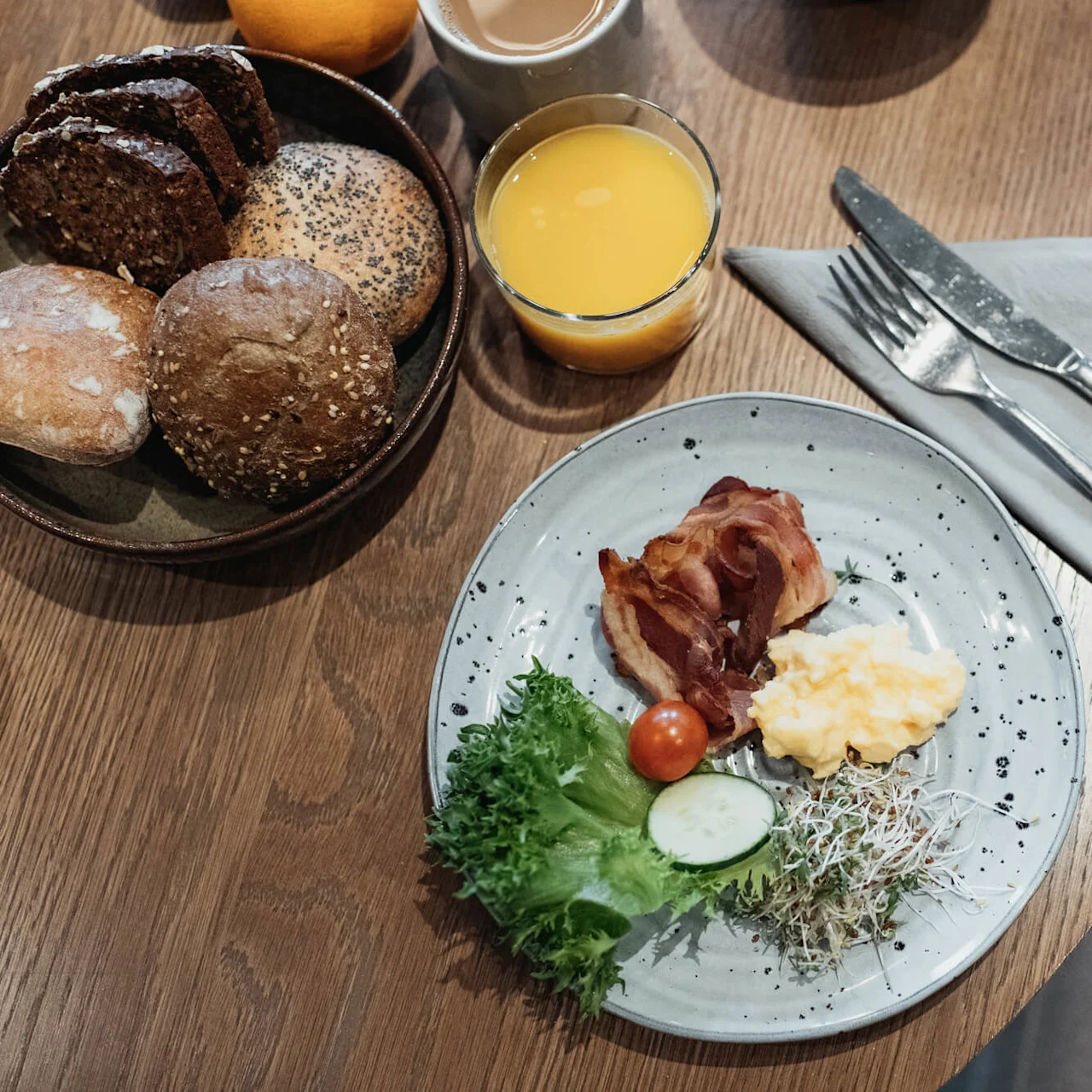 A breakfast plate with bacon, scrambled eggs, and salad, served with assorted breads and orange juice on a wooden table.