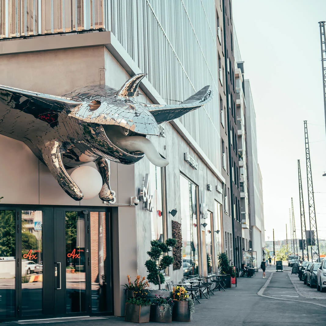 A large, shiny manta ray sculpture protruding from a building facade above a doorway, with cafe tables nearby on a city street. Text: "Ax" and "AVEQIA".