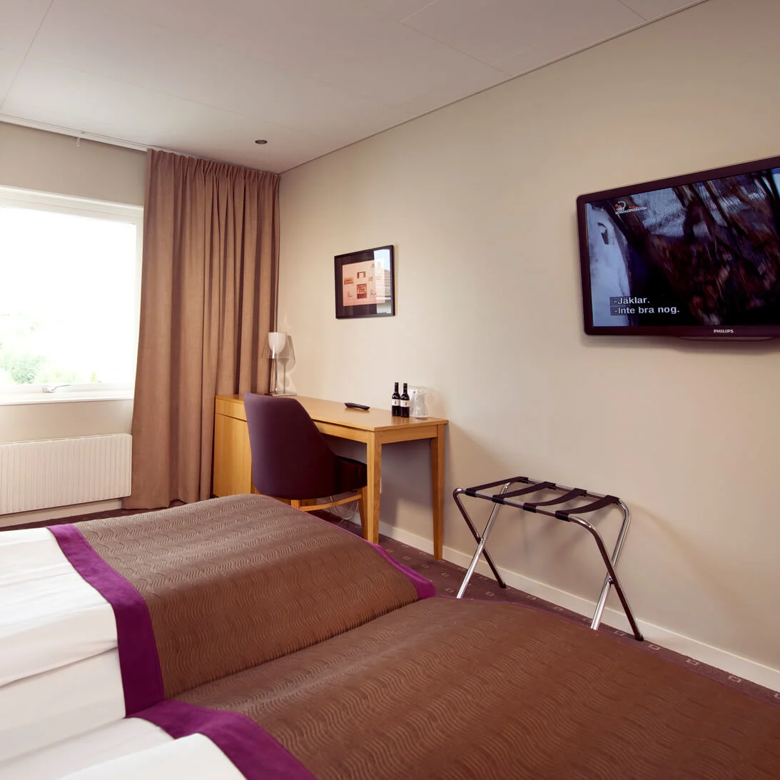 A hotel room with a bed, armchair, desk, and mounted television displaying a scene with subtitles: "...skakar lite bra nog."