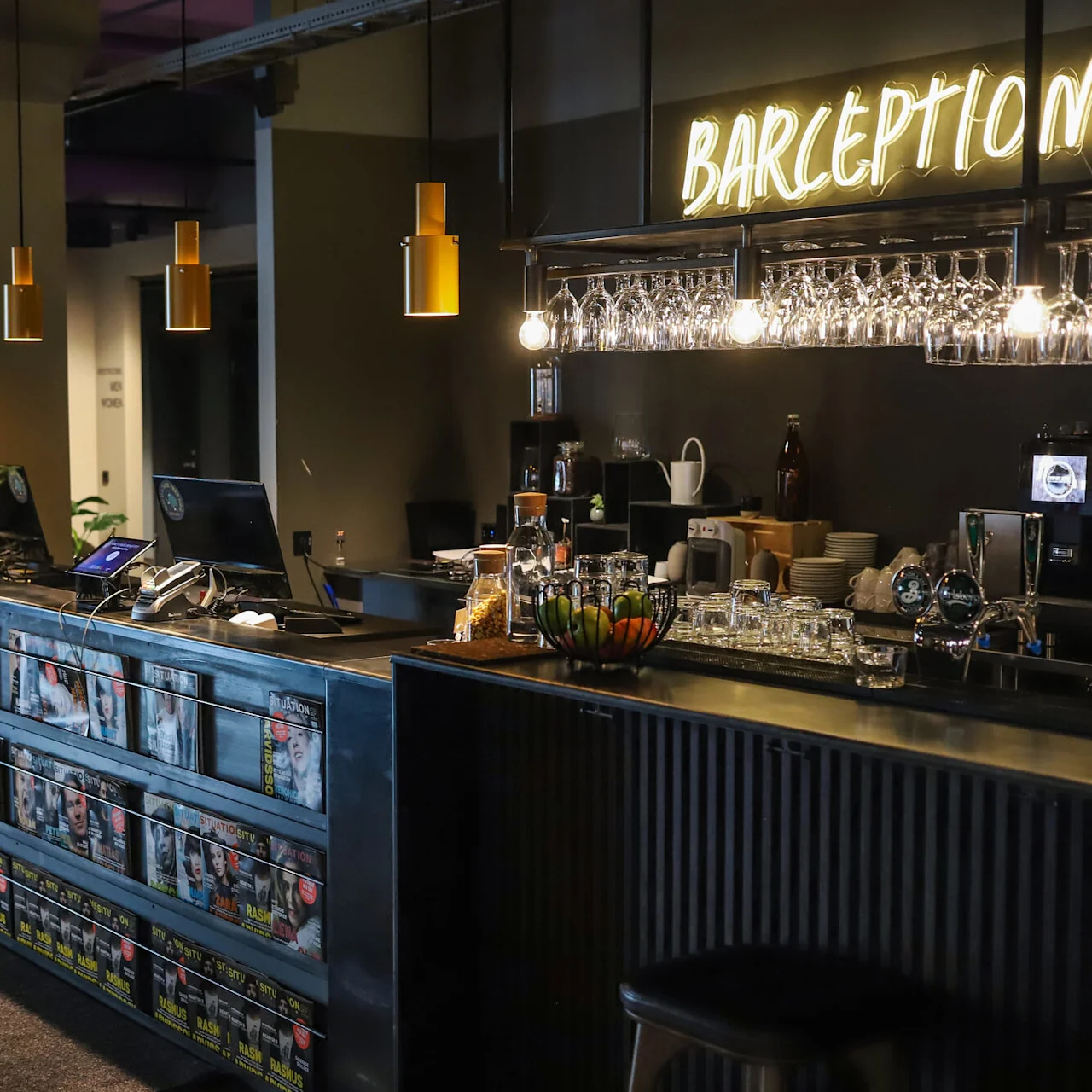 A bar counter doubling as a reception desk with pendant lights, in a dimly lit, modern space with shelves of snacks.