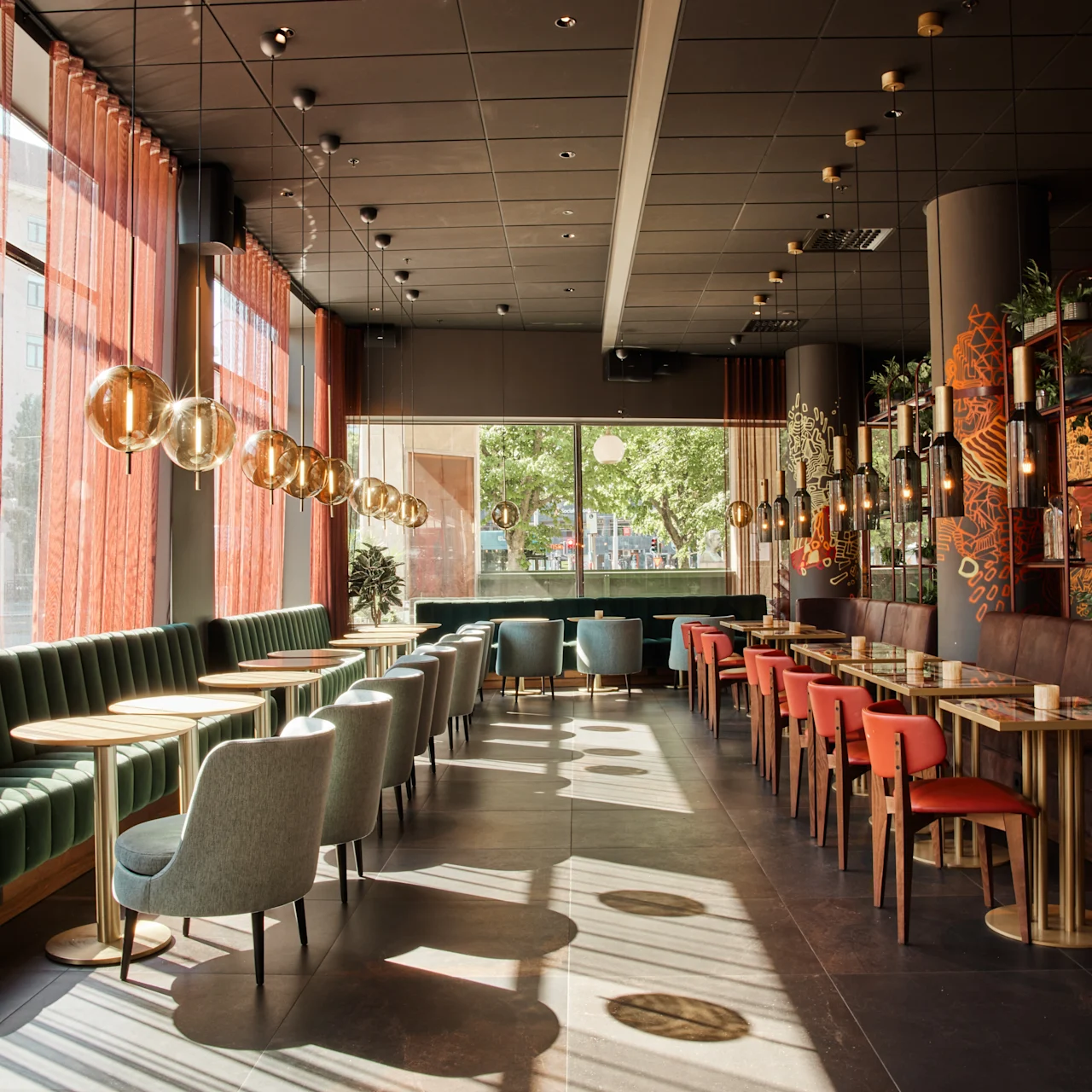 Modernt café med trämöbler och färgglada stolar prydligt arrangerade. Solljus strömmar genom stora fönster med skira gardiner. Dekorativa hängande ljus skapar värme mot en bakgrund av växter och livfulla väggkonst.