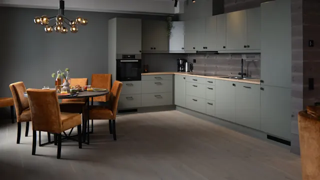 Modern kitchen with light green cabinets and wooden countertops, featuring a round dining table with brown chairs. Overhead, a stylish chandelier hangs. The floor is light grey wood.
