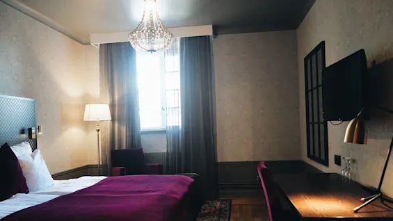 En seng med lilla tæppe står ved et vindue, omgivet af en lampe, stol og skrivebord i et varmt oplyst værelse.