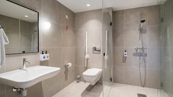 A modern, accessible bathroom with light grey tiles, featuring a sink, toilet, and a walk-in shower with grab bars.