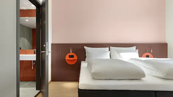 Et moderne hotellrom med en komfortabel seng, en rosa vegg og et glimt inn i et stilfullt flislagt bad.