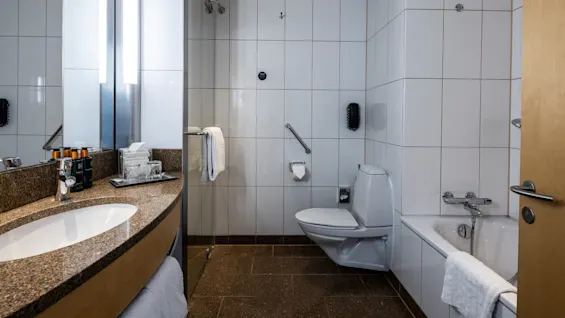 Ett badrum med ett handfat av granit, toalettartiklar och handduk till vänster, en toalett i mitten och ett badkar med en handduk till höger. Väggar är kaklade vita.