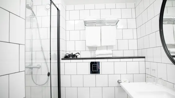 En bruser med glasdøre er i et hvidt flisebelagt badeværelse. To foldede håndklæder ligger på en væghylde over et toilet. Et spejl og vask til højre.