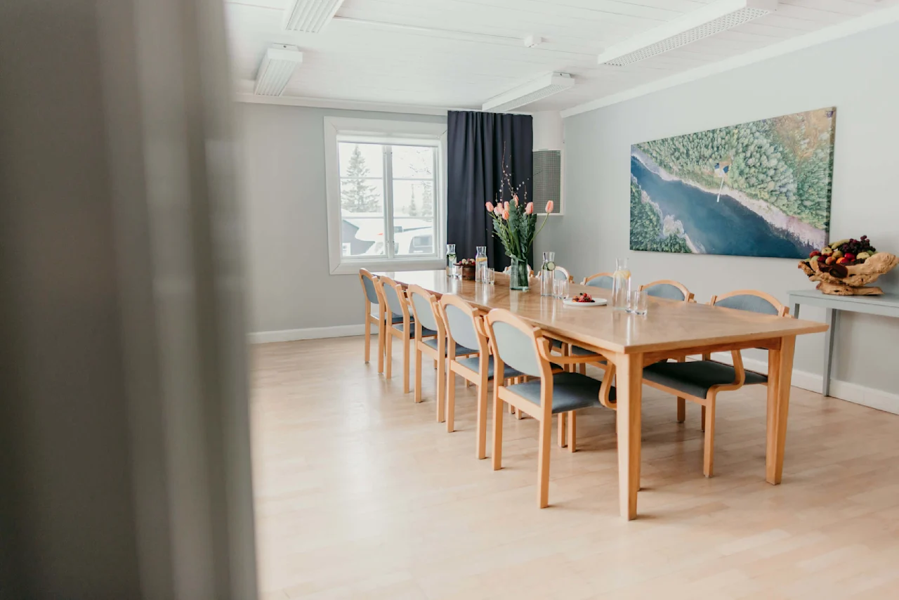 Et træspisebord med stole, dækket med klare glas, blomster og frugt, i et lyst, moderne rum med et stort landskabsbillede på væggen.