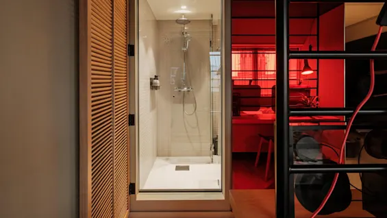 Shower with transparent glass door stands in a small, well-lit bathroom. Adjacent is a cozy room with red lighting, visible through an open doorway.