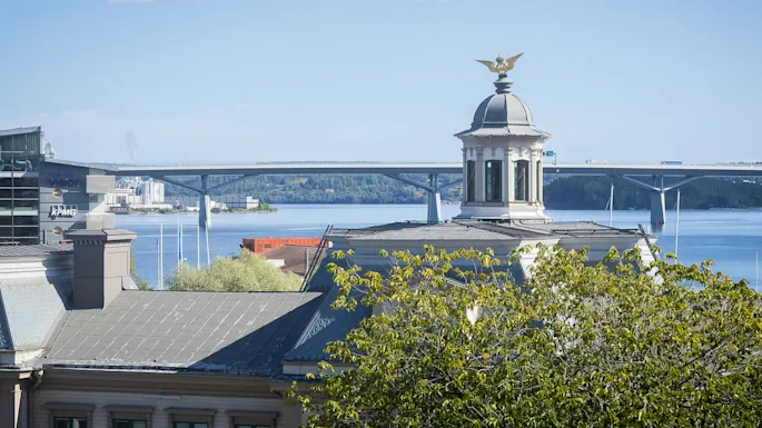 En klassisk bygningskuppel overser en moderne bro som strekker seg over et vann med trær og skyskrapere, under en klarblå himmel.