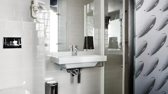 A clean, modern bathroom features a white sink with a mirror above and a chrome faucet. It includes tiled walls, a shower, a small trash can, and a hairdryer.