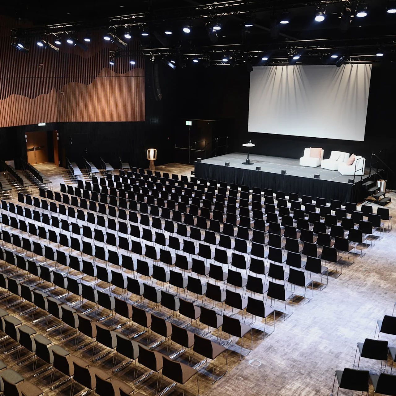 En tom sal har pent arrangerte svarte stoler vendt mot en scene med sofaer og en projektorskjerm, under sterkt overhead lys, i et trepanelt rom.