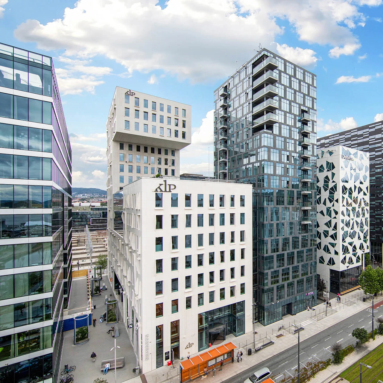 Tall, modern buildings stand along a street with pedestrians and cyclists. "KLP" and "Deloitte" are visible on two buildings. Trees line a tramway under a partly cloudy sky.