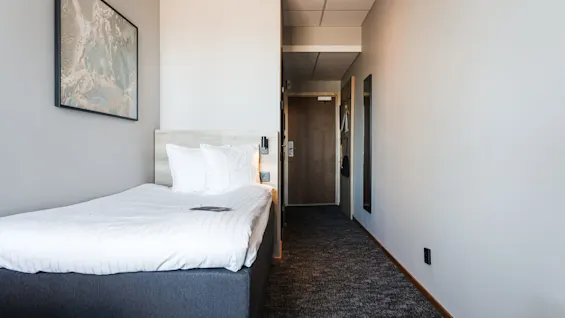 En prydligt bäddad säng står i ett smalt hotellrum, med ett trähuvudgavel och en abstrakt målning på väggen ovanför. Gången leder till en stängd trädörr.