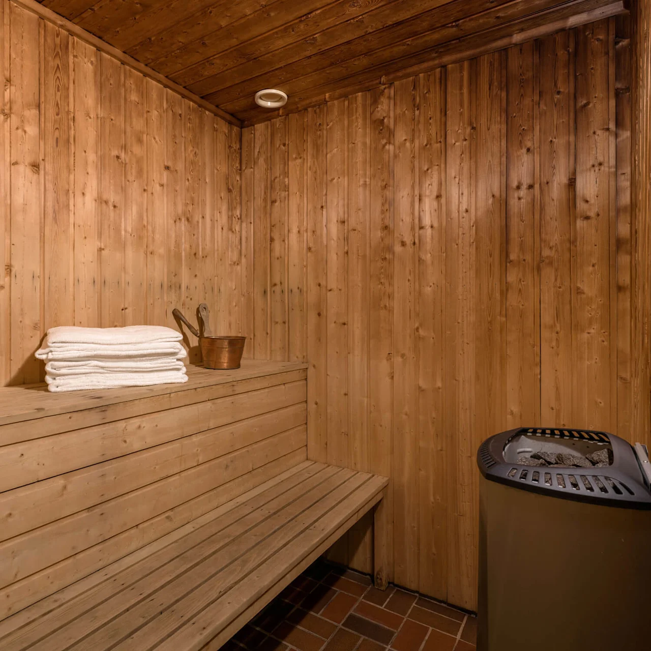 A wooden sauna bench with stacked white towels and a wooden bucket sits next to a metal stove on a tiled floor, surrounded by wooden-paneled walls.