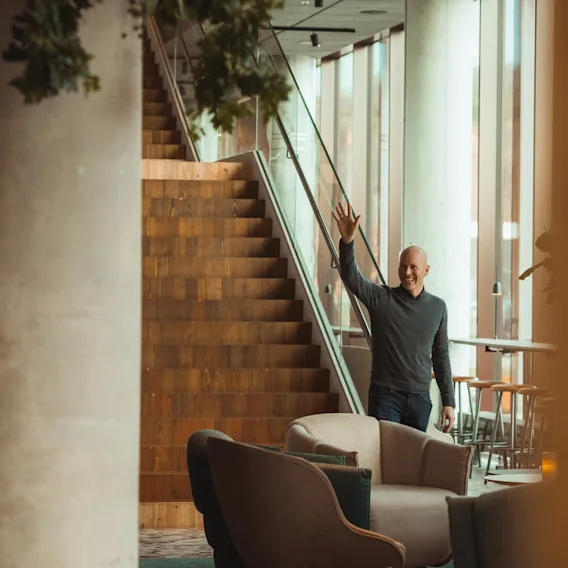 En person vinkar glatt, stående nära en trätrappa inuti en modern byggnad med stora fönster, omgiven av bekväma sittplatser och grönska.