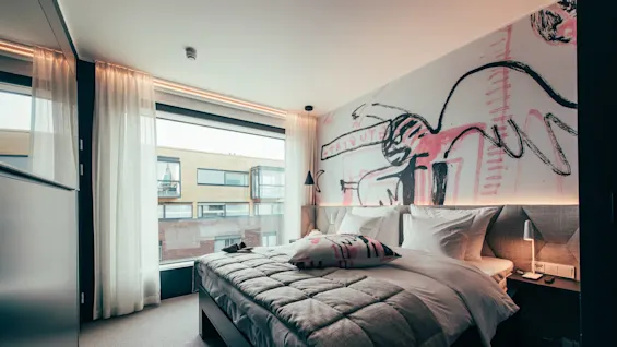 A neatly arranged bed with patterned pillows and quilt sits against an artistic wall in a modern room with large windows and soft curtains, overlooking adjacent buildings.