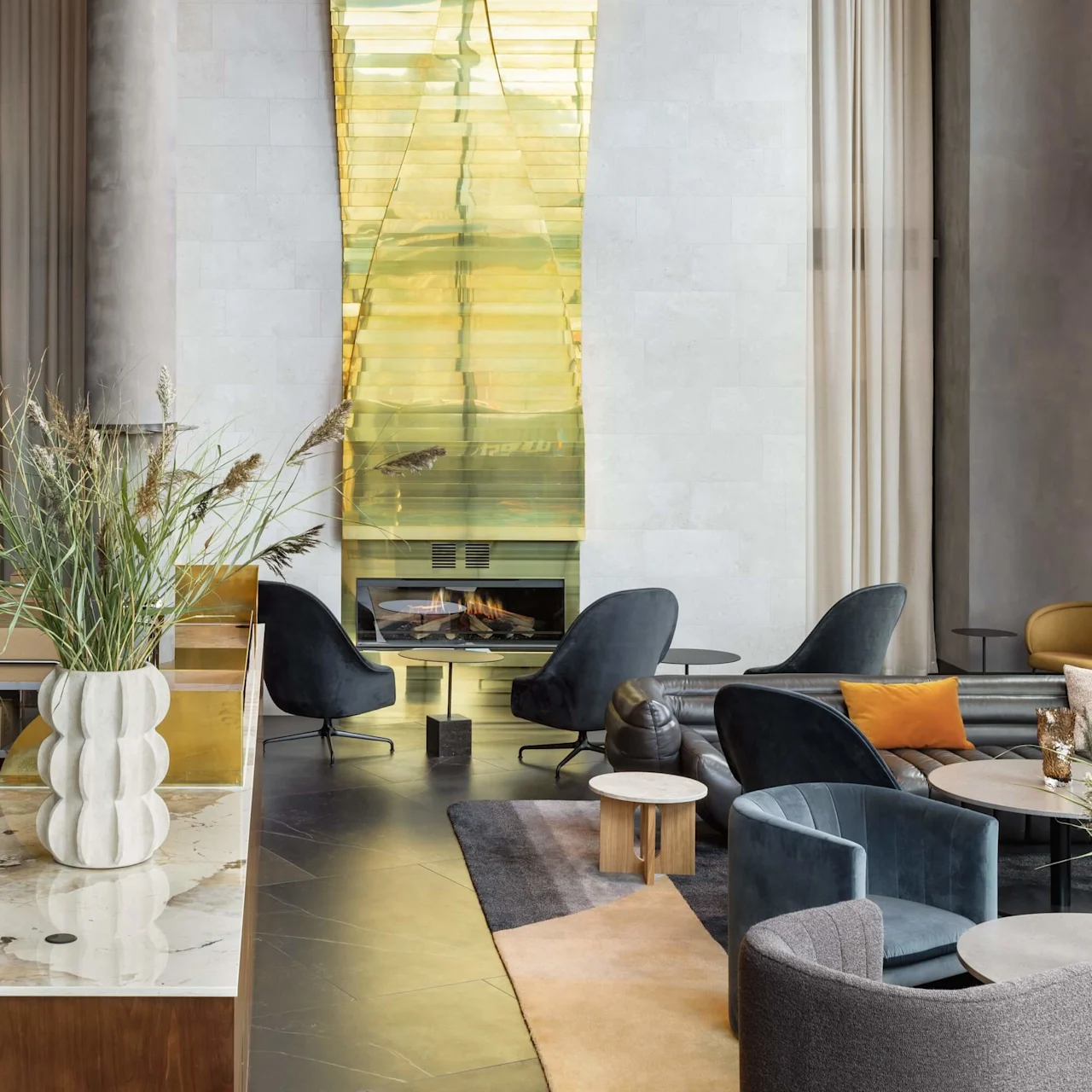 En modern lounge med blå stolar arrangerade runt ett lågt bord, mot en eldstad med gulddetaljer på en texturerad vägg, med hög takhöjd och minimalistisk inredning.