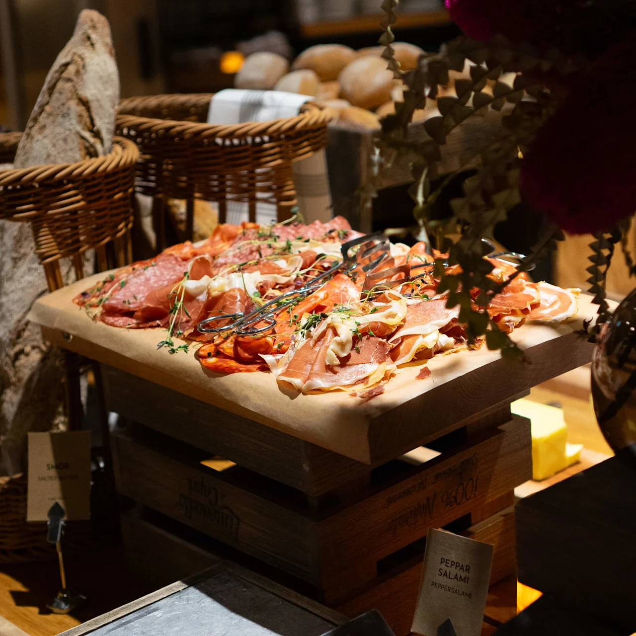 Kurerte kjøtt på bakepapir oppå trekasser, omgitt av brødkurver og grønt i rustikk miljø. Skiltet lyder: "PEPPARSALAMI PEPPERSALAMI".