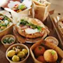 Erilaisia italialaisia ruokia, kuten pizzaa, salaattia, voileipiä ja oliiveja, on asetettu puupöydälle, mikä viittaa rentoon ruokailu- tai takeout-tilanteeseen.