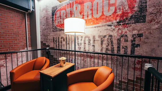 Två orange stolar placerade runt ett litet bord med popcorn, under en hängande lampa. Bakom finns en texturerad vägg med texten "ART & ROCK HIGH VOLTAGE" i en industriell loftstil.