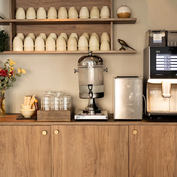 En självbetjäningsstation med kaffemaskin som erbjuder dryckesval, en vattendispenser och koppar. PAPPERSKORG.