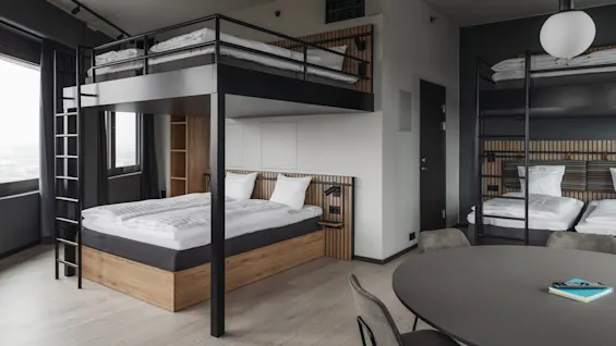 A modern hotel room features a double bed with a lofted bunk above, surrounded by wood accents and large windows. A round table with chairs is in the foreground.