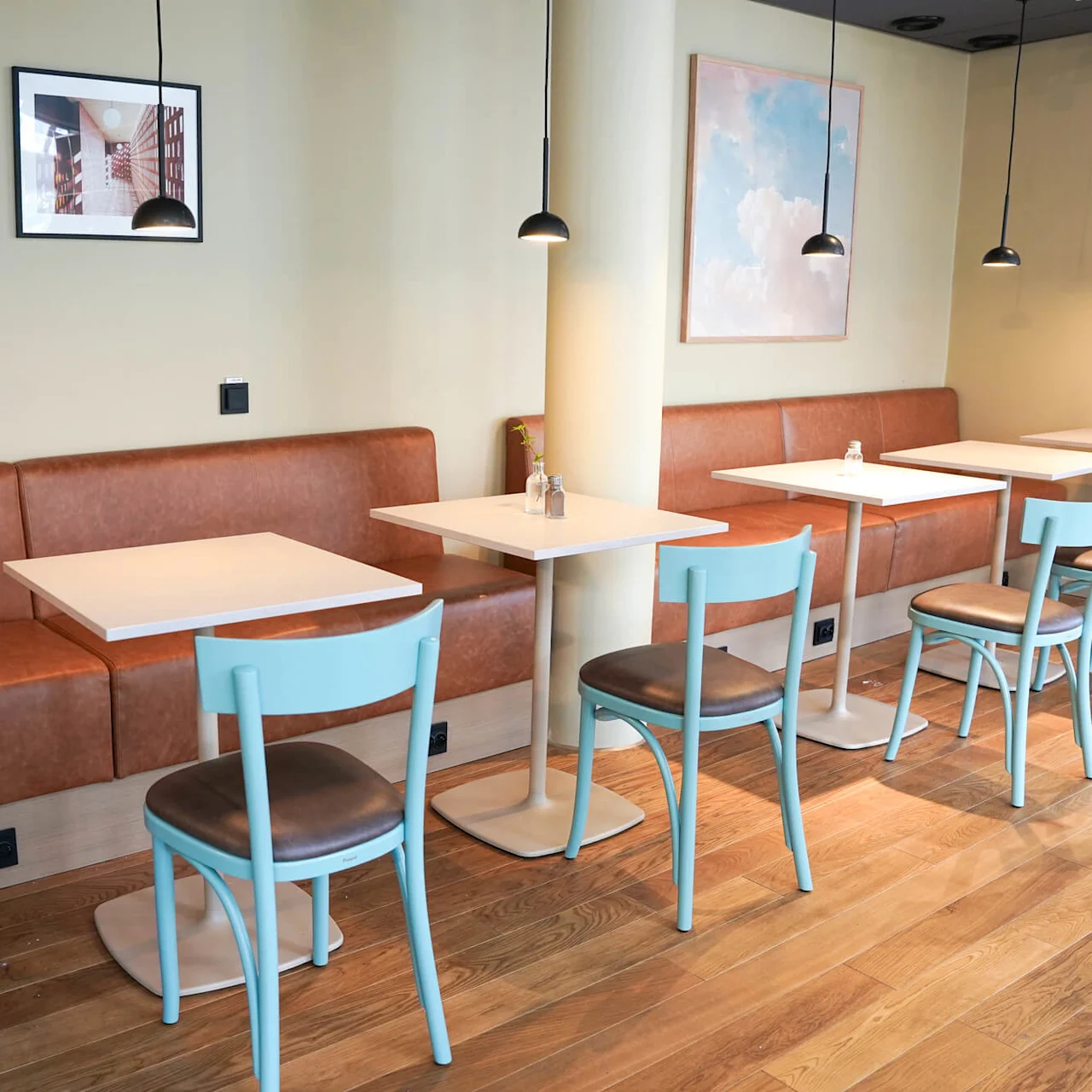 Café interior features light blue chairs around white tables, with brown leather booth seating. Framed artwork and hanging pendant lights create a cozy ambiance on wooden flooring.