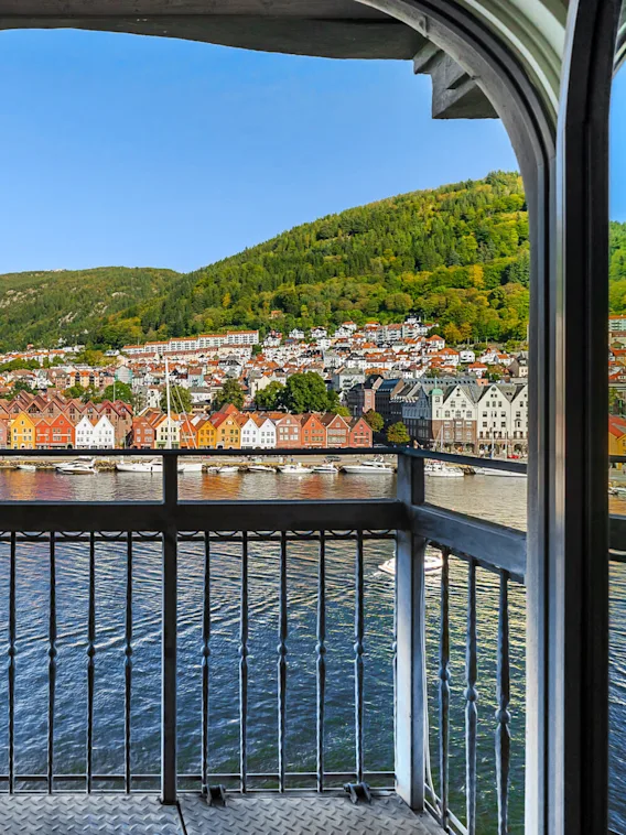 A balcony overlooks a harbor with colorful houses along the waterfront. Mountains rise in the background under a clear blue sky.