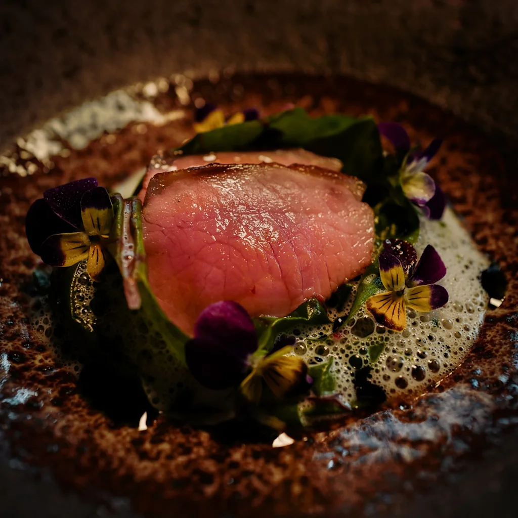 Et nærbillede af en smukt anrettet ret med mørt kød, levende spiselige blomster og en delikat skumsauce.