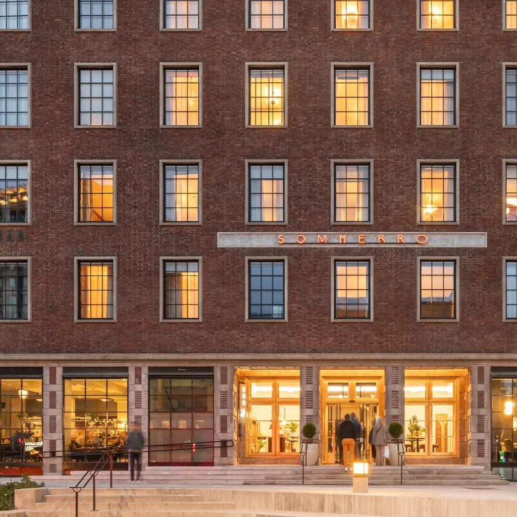 En tegelbyggnad med upplysta fönster, människor går in genom dess ljust upplysta entré. Skylten ovanför lyder "SOMMERRO." Omgivningen har trappor och planterade buskar.