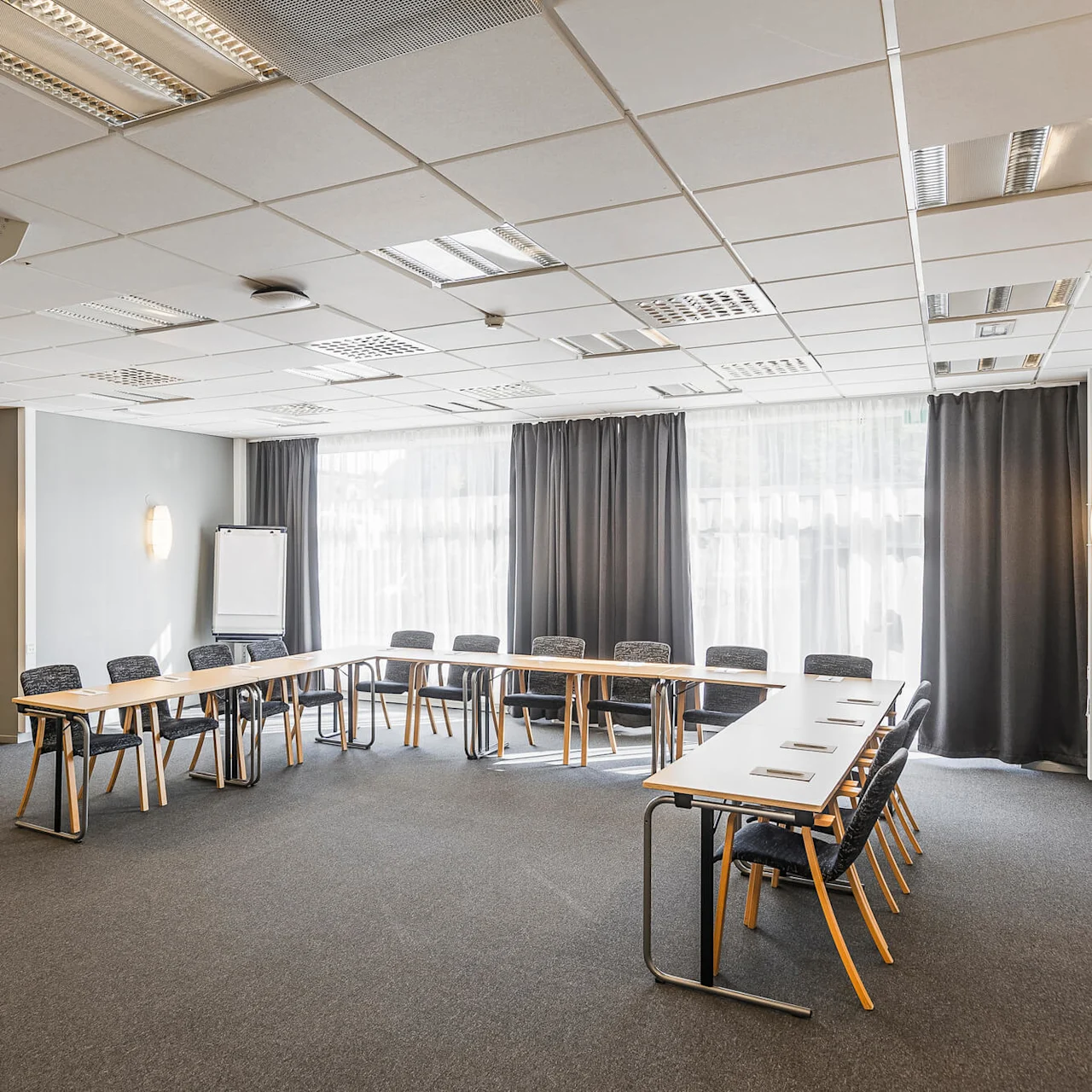 Tomt konferensrum med bord i U-form, stolar runtomkring, en projektor i taket och en whiteboard, i en välbelyst inredning.
