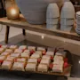 Små kakrutor med vit glasyr och jordgubbar ligger på en bricka. De står på en diskbänk under en hylla med staplade tallrikar och ett ljus.