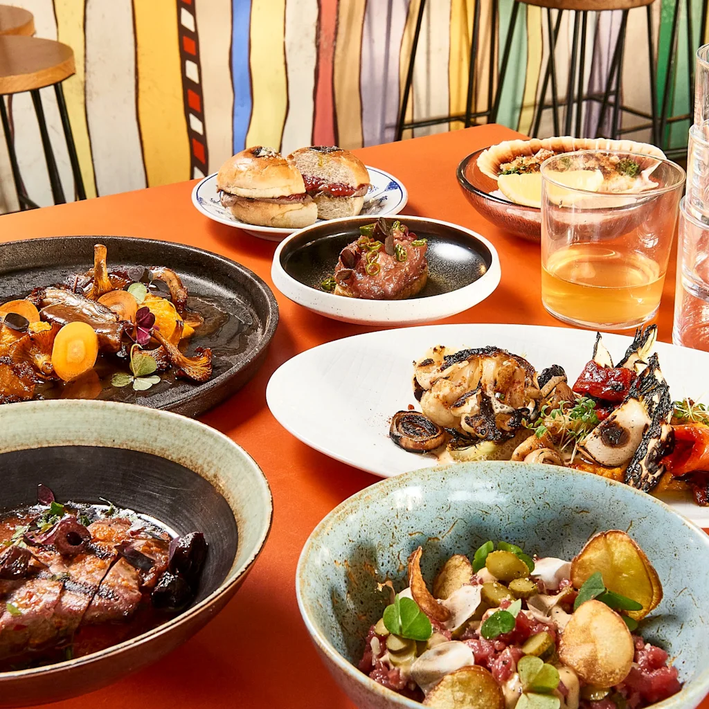 Eloisa pöytä katettuna herkullisilla ruokalajeilla, kuten lihalla, kasviksilla ja pienillä hampurilaisilla, sekä juomilla.