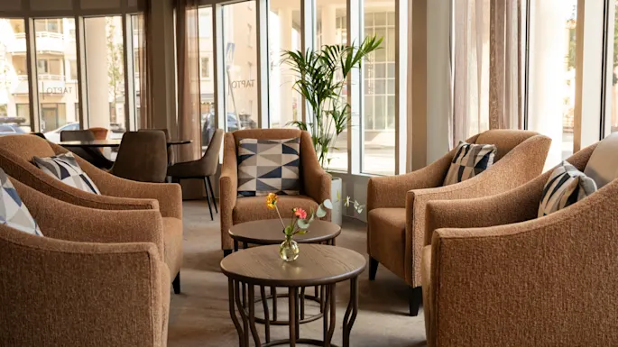 A cozy hotel lounge with comfortable armchairs and small tables, bathed in natural light from large windows.