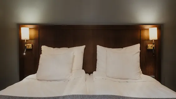 A cozy hotel bed with white linens, a wooden headboard, and warm wall lighting, inviting rest at Home Hotel Kung Oscar.