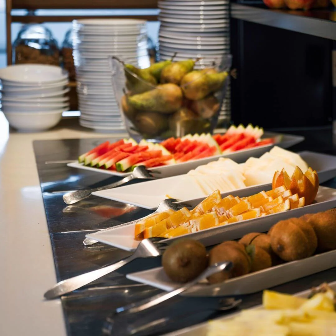 Frugtfade med kiwi, vandmelon og appelsinskiver er arrangeret på en buffetbord. I baggrunden er der skåle, stablede tallerkener, krukker og yoghurtpakker mærket "Alpro" og "Yoghurt Uten."