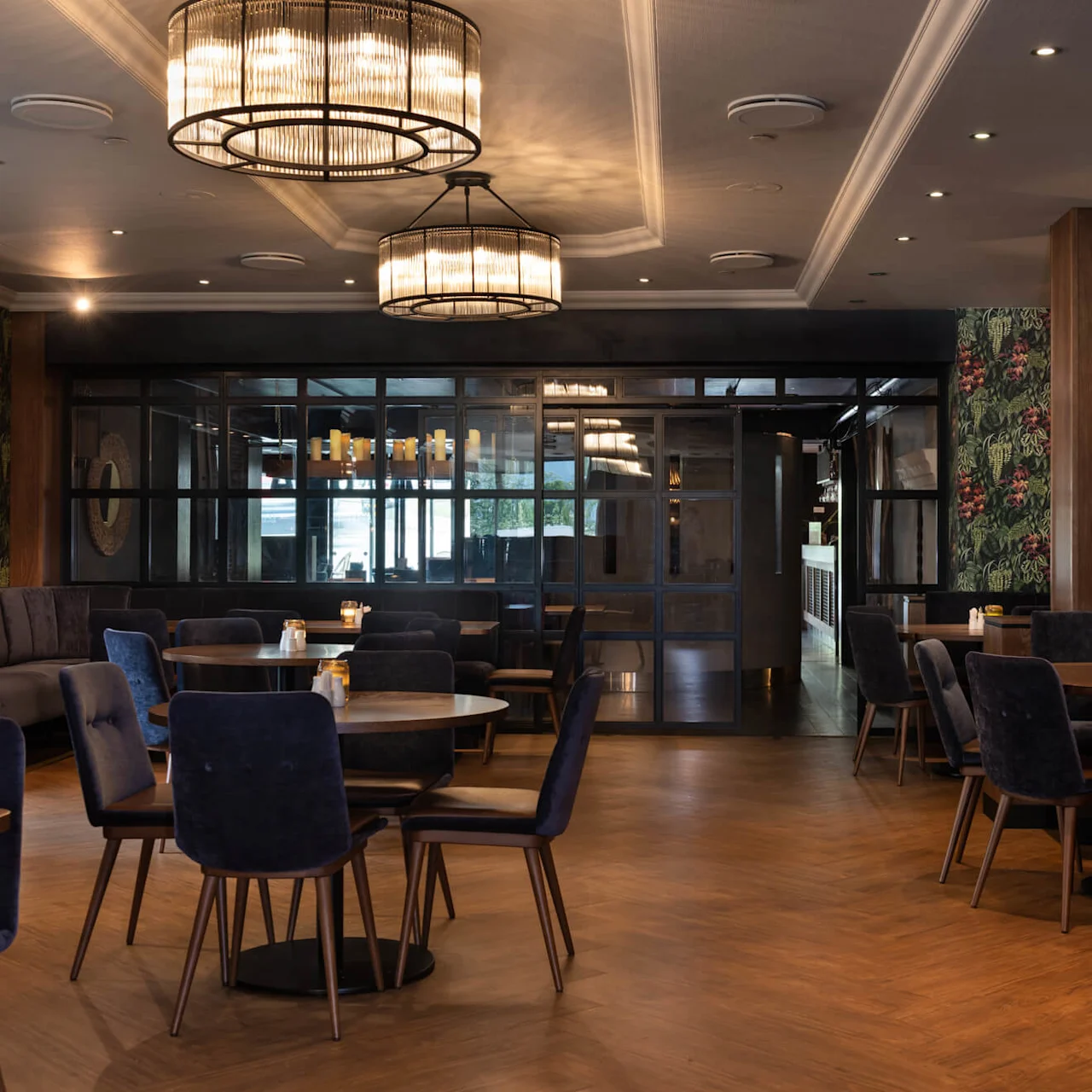 En mysig restaurang har runda bord och blå stolar. Stora taklampor lyser upp ett trägolv och tapeter med gröna mönster. Den svagt upplysta atmosfären förstärker den eleganta, moderna miljön.