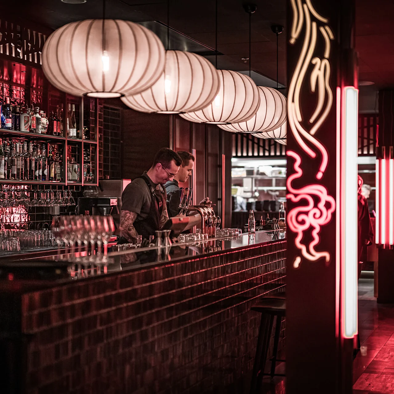 Bartendrar förbereder drinkar i en svagt upplyst bar med hyllor av flaskor och glas. Stora hänglampor hänger ovanför och neonlys skapar en röd stämning.