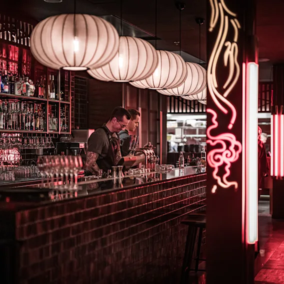 Bartendere tilbereder drinker i en svakt opplyst bar med hyller av flasker og glass. Store hengelamper henger over og neonlys gir en rød stemning.