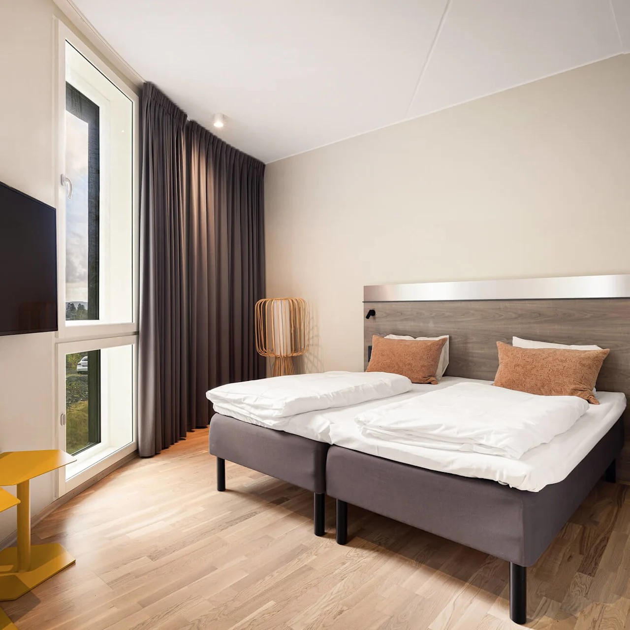 A neatly made bed with white and brown pillows stands in a bright room with light wooden floors, a wall-mounted television, and large window with dark curtains.