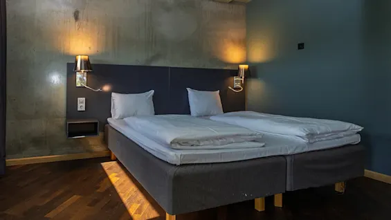 A bed with white linens is positioned in a dimly lit room. Wall-mounted lamps provide soft lighting. The wooden floor adds warmth to the modern, minimalist design.