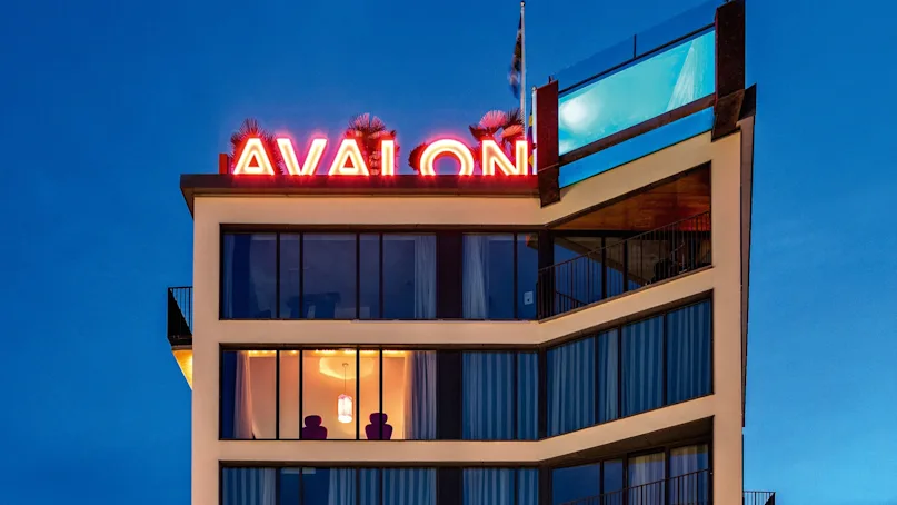 A modern multi-story building with large windows displays "AVALON" in red neon. Two people stand on a balcony. Below, a lit restaurant bar sits in a quiet street.