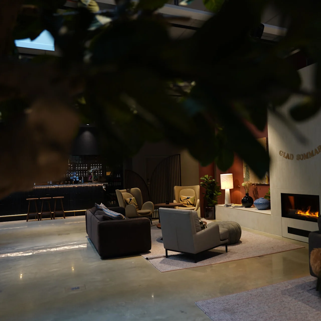 A cozy living room with a lit fireplace that reads "GLAD SOMMAR", modern furniture, and a bar in the background.