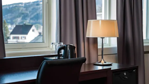 En skrivebordslampe oplyser et træskrivebord med en elkedel og glas, mod vinduer med bjergudsigt.