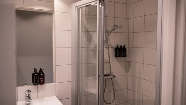 Dusch innesluten av glasdörrar, med ett modernt duschmunstycke och tre svarta flaskor fästa på vita kakelväggar. Bredvid en vit handfat med kromkran och matchande flaskor under en spegel.