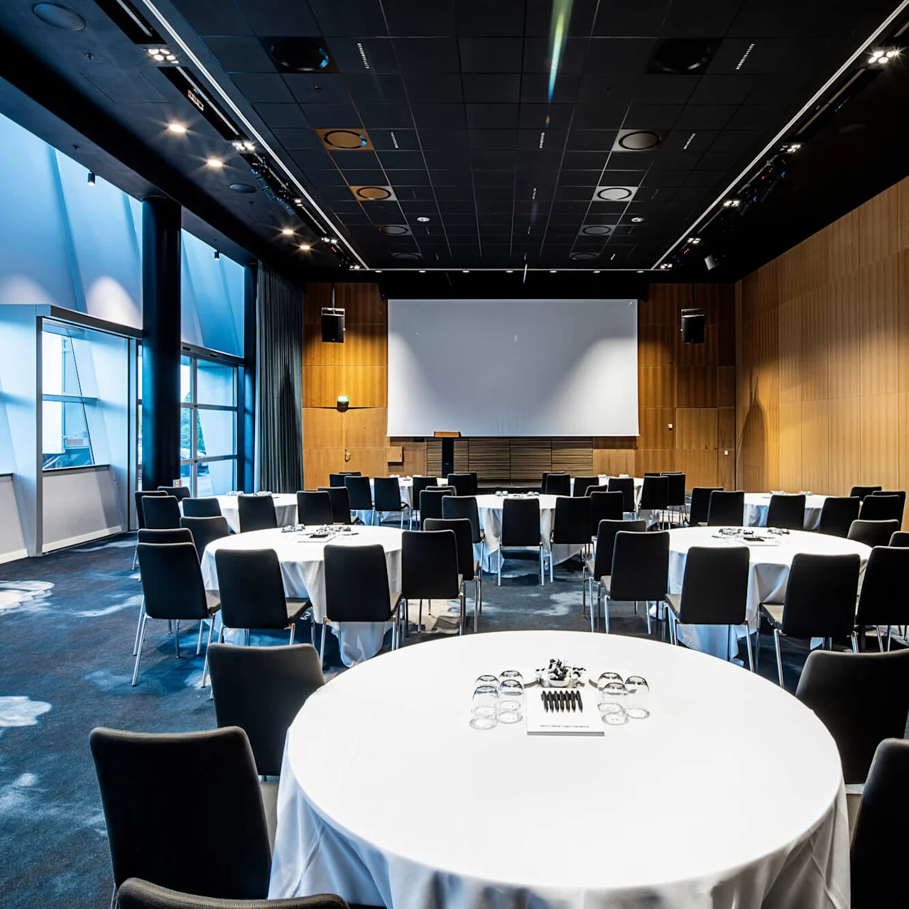 Konferensrum med runda bord uppställda för möte, med svarta stolar, vita dukar och stor projektorduk. Stora fönster släpper in naturligt ljus i modern miljö.