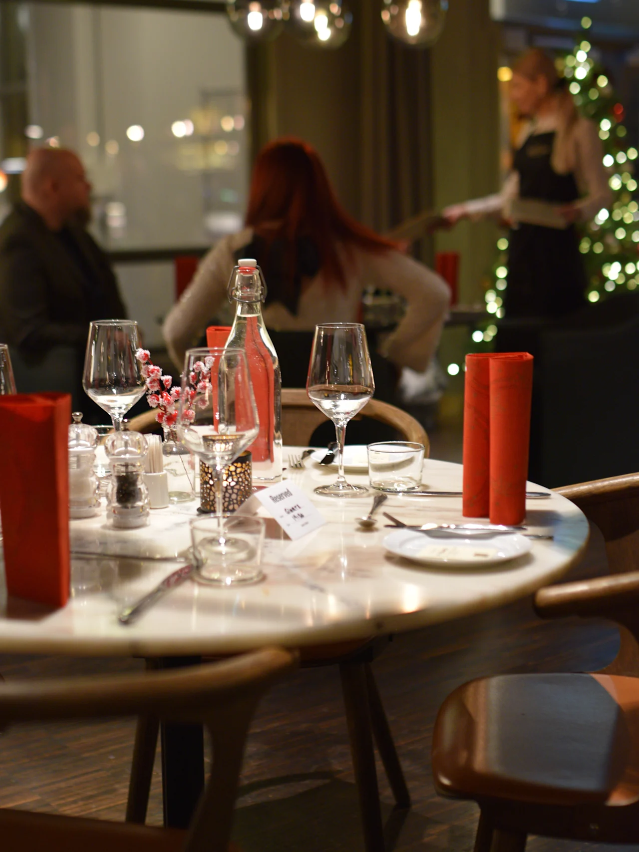 A round table set with glassware, a water bottle, and folded red napkins in a cozy restaurant. People converse in the background near a Christmas tree.